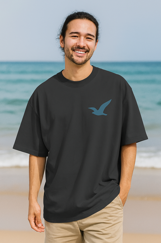 Man wearing a black t-shirt with a blue bird logo on a beach